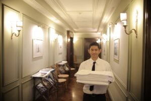 A young male hotel staff member carrying fresh towels in a beautifully lit hotel hallway.